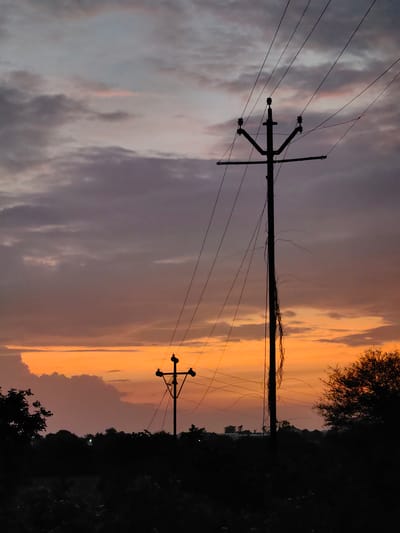 This is a silhouetted image of a sunset in Dattapur, India, with two utility poles and power lines prominently featured against a dramatic sky. The sky displays a gradient of colors, transitioning from a vibrant orange and yellow near the horizon to muted purples and grays higher up, with scattered clouds. The foreground is dark and obscured, showing the silhouettes of trees and vegetation. The utility poles are tall and black, with wires extending between them and across the frame. One pole is closer and larger, while another is smaller and further in the distance. There are no people or animals visible in the scene. The overall impression is one of a rural or semi-rural landscape at dusk.