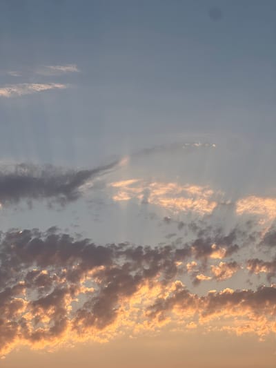 The image displays a sky with scattered clouds, tinged with the warm colors of sunrise or sunset. The clouds vary in density and color, with some appearing dark and ominous, while others are illuminated with golden and pink hues. The sky itself transitions from a pale blue at the top to a softer, warmer tone near the horizon. There are no visible people, subjects, objects, actions, or interactions depicted in the image. The scene is solely focused on the natural phenomenon of the sky and clouds. It is not possible to determine the specific location in the city of Rotmanka, Poland from the provided image.