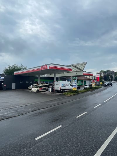 This is a medium-distance, eye-level, outdoor, over-the-shoulder shot of a gas station on a cloudy day in Lauterbach, Germany. The gas station is identified by the "AVIA" logo. Several vehicles are present, including two white taxis, a white van labeled "Hermes", a dark SUV, and a black sedan. The ground appears wet, suggesting recent rain. The sky is overcast with heavy clouds. The architecture of the gas station includes a covered area with red and white trim, and a separate building with a dark facade. A price board displaying fuel prices and advertisements for services like "GREENBOX", "AdBlue", and "LOTTO" are visible. Buildings and trees are present in the background. The road in the foreground is asphalt and wet, with white lane markings.
