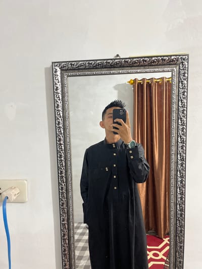 An individual is depicted taking a mirror selfie indoors in Alueduamuka S, Indonesia. The person, whose face is largely obscured by a dark-colored smartphone held in their right hand, has short, dark, spiked hair. They are wearing a full-length black garment featuring a mandarin collar and approximately eight light-colored buttons extending vertically down the front. A wristwatch with a dark face and a green bezel is visible on their left wrist, with two light-colored buttons present on the cuff of the left sleeve.

The selfie is captured via a large, rectangular mirror featuring an ornate, silver-colored frame. The background reflected in the mirror includes a set of brown, pleated curtains with gold-colored eyelets and a decorative finial on the right. Below the curtains and the individual, a patterned surface is visible, consisting of a white and grey checkered pattern on the left and a red and white patterned rug or mat on the right. Outside the mirror's reflection, on the immediate left, a light-colored wall contains a white electrical outlet with two sockets. A blue cable is plugged into the upper socket and extends downwards.
