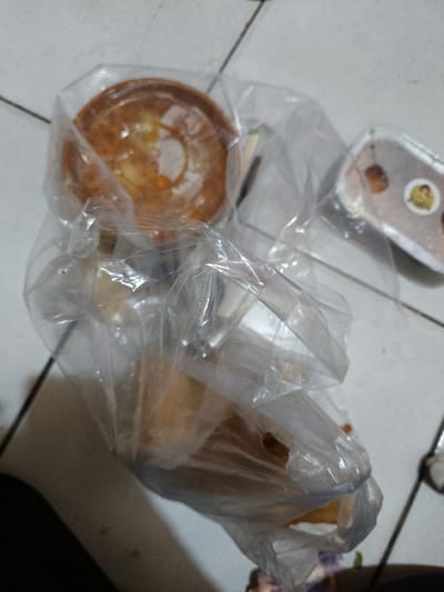 In Cileungsi, Indonesia, a transparent plastic bag containing multiple food items rests on a light-colored tiled floor with dark grout lines. Prominently visible within the bag is a round, golden-brown baked item, possibly a cake or pie, showing a textured top. Other lighter-colored food items, consistent with pastries or bread, are also contained within the bag. To the right, a separate clear plastic container with a white base holds dark-colored contents and features a circular sticker on its lid. No human figures or active interactions are visible in the scene.