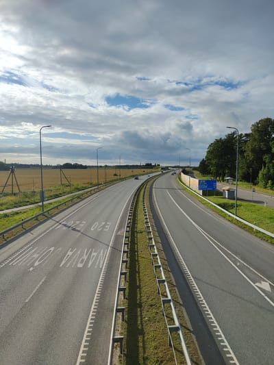The image shows a highway in Edise, Estonia, with a cloudy sky. The highway is divided into two directions by a guardrail and grass. There are streetlights and road signs visible. The road has lane markings and some text painted on it, reading "NARVA" and "E20". There are a few cars on the road and some trees and fields on the side. There are also some small structures that look like bus stops.