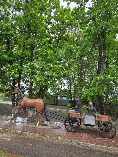 This image depicts a sculptural tableau in Valdgeym, Russia, featuring a horse-drawn cart with two human figures. The scene is set on a mixed ground surface comprising dark asphalt on the left and reddish-brown brick paving on the right, which shows signs of wetness.

On the right side of the paving, a brown horse statue with a dark mane and tail, and white lower legs, is positioned with its front left leg lifted. Black shafts extend from the horse to a two-wheeled wooden-textured cart, suggesting the horse is pulling it. Seated in the cart are two human figures: a male figure in the front and a female figure behind him. The male figure is dressed in dark clothing, including a cap, and has a beard. The female figure is dressed in dark clothing with a head covering, resembling a scarf. Both human figures and the cart appear to be made of a solid, painted material, possibly concrete or wood.

Behind the statues and paving, a row of large, deciduous trees with dense green foliage forms a significant part of the background, obscuring the sky. Their trunks are visible between the leaves. A dark green corrugated metal fence runs horizontally through the midground, separating the immediate area from a light blue-green wooden house with white window frames visible on the far left. The ground under the trees and near the fence is covered in dark earth and some sparse vegetation. Scattered green leaves are present on the wet brick paving. The overall lighting suggests an overcast day.