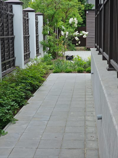 This image captures an outdoor scene featuring a paved pathway, lush garden, and various architectural elements, set in Березка Снт (красногорский Р-н), Russia.
In the foreground, on the left, a multi-tiered white building or fence structure with dark brown, lattice-like panels is partially visible, with white wires draped over some sections. Overhanging this structure is dense green foliage from trees and bushes.
Adjacent to this, running through the mid-ground, is a vibrant garden bed teeming with various green plants and shrubs. A prominent feature within the garden is a bush bearing numerous white flowers, likely roses. A black plastic crate is partially visible among the plants further back in the garden.
To the right of the garden, a light grey paved walkway, constructed from square tiles, stretches into the distance. Above the walkway, a concrete overhang or ceiling features small, grey cylindrical protrusions, possibly lighting fixtures or sensors.
In the background, on the left, a building with brown vertical siding is visible through the trees. Above this, a dark brown wooden pergola or trellis structure with horizontal beams frames the top of the scene.
The scene depicts a static outdoor environment under natural, diffused daylight, suggesting either an overcast day or a shaded area, as there are no strong shadows. No people or visible text are present. The overall impression is one of a well-tended garden and pathway within a residential or cultivated area.