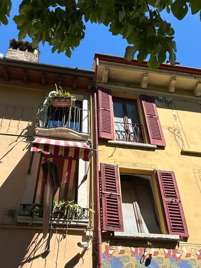 This is a low-angle, medium shot of a yellow building in Desenzano del Garda, Italy, on a bright, sunny day. The upper part of the building features an ornate cornice supported by corbels, and a dark red roof. A chimney made of brick and stone is visible to the left. Green leaves from a tree canopy hang over the scene, casting shadows.

The building has several windows. The left side shows a balcony with a railing and a potted plant. Below the balcony, a red and white striped awning covers a door. A security camera is mounted on the wall below this window.

To the right, two windows are visible. Both have open, dark red shutters. The upper window has a small balcony with a railing. The wall of the building is decorated with a light-colored, Art Nouveau-inspired mural featuring floral motifs. The lower window also displays floral decorations below its sill.

The overall impression is one of a charming, sun-drenched European building.