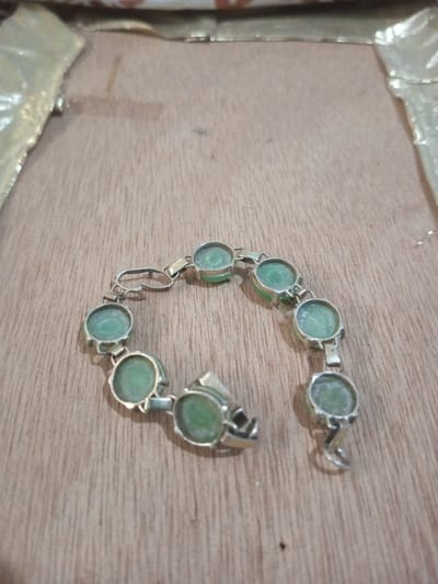 The image presents a metallic link bracelet, exhibiting ten circular, opaque, light green stones, positioned on a light brown wooden surface. Each stone is encased within a metallic bezel, with the bezels connected by small, flat, rectangular metallic links. The bracelet is unfastened; a hook-style clasp is visible at the left end, and a corresponding loop at the right end. The metallic components display a silver-toned, possibly aged, appearance with visible wear and surface irregularities. The green stones appear somewhat dull or dusty. The wooden surface beneath the bracelet features a horizontal grain pattern and minor abrasions. In the blurred upper background, two crumpled, gold-toned elements are partially visible, suggesting a distant background or packaging material. No individuals are present, and no active interactions or actions are depicted. The scene is located in the city of Chandragiri, Nepal.