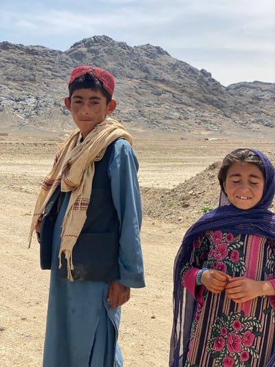 Two children stand outdoors in a dry, mountainous landscape. On the left is a boy wearing a red cap, a light-colored scarf, a blue tunic, and a dark vest. On the right, a girl wears a blue headscarf and a striped dress with floral patterns. The background features a mountain range under a partly cloudy sky. The ground is dry and appears arid.