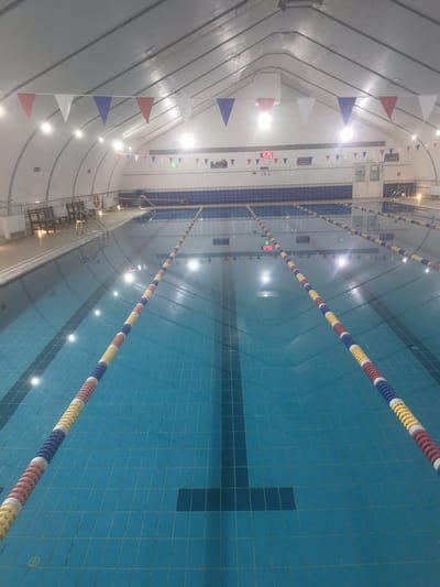 This media file depicts an indoor swimming pool in Қосшағыл ауылдық округі, Kazakhstan. The scene is dominated by the large, blue swimming pool with marked lanes. Several lane ropes, colored red, white, yellow, and blue, extend the length of the pool, demarcating the swimming lanes. The pool is empty, suggesting no activity or event is currently taking place.

The setting is an indoor facility with a curved ceiling structure. Along the top of the pool area, strings of red, white, and blue triangular flags are hung, adding a festive touch. Lights are visible along the ceiling, illuminating the space. There are benches and railings along the side of the pool.

A digital clock is visible on the wall above the pool, displaying the time "15:20". Some signs with text in Cyrillic script are also present on the walls, including "НЕ БЕГАТЬ" which translates to "DO NOT RUN". The overall atmosphere is neutral and calm, with no visible people or explicit emotional cues. The lighting suggests it is likely daytime or evening, with artificial lighting being the primary source. The presence of the flags and the clean, organized appearance of the pool indicate it is likely a public or community facility.
