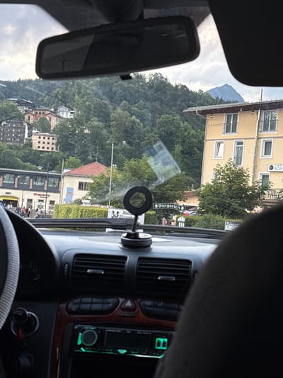 The image shows a view from inside a car looking out at the town of Berchtesgaden, Germany. In the foreground, the car's dashboard is visible, including the steering wheel, air vents, and a car stereo with green illumination. A circular object, likely a phone mount, is attached to the windshield.
Outside the car, a street scene unfolds. To the left, a building with shops is visible, with signs indicating "Gesundheit" (Health) and "Shape Up". A price of "34,90 € / Monat" (34.90 € / Month) is also displayed. People are walking on the sidewalk in front of the shops. Further down the street, a building with a red roof and a Bavarian flag emblem is present, with trees and greenery surrounding it.
To the right, a yellow building, identified as "Hotel Schwabenwirt" by a sign, stands prominently. This building has multiple windows and balconies. Above the hotel and the town, a lush green mountainside covered in trees rises, with a distinct mountain peak visible in the distance under a cloudy sky. A sign for "Biergarten" (Beer Garden) is also visible. The overall impression is a scenic view of a European town nestled in a mountainous region.