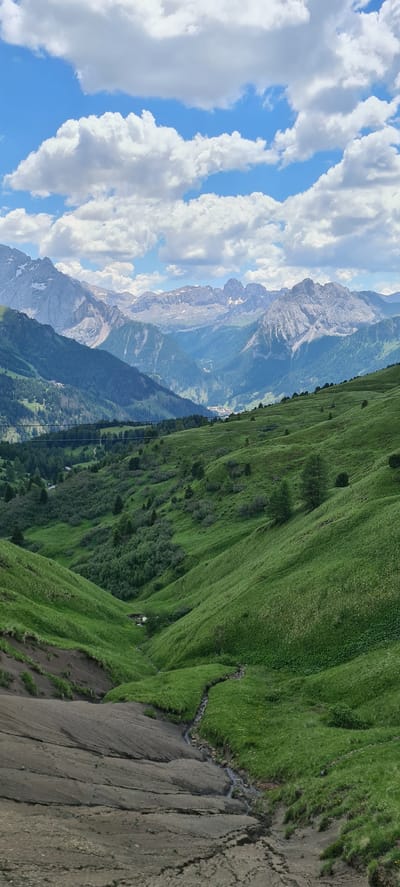 The image depicts a vast, verdant mountain landscape under a partially cloudy sky. The foreground features a steep, rocky, and barren slope with visible erosion marks, transitioning into rolling green hills covered in grass and scattered trees. A narrow stream meanders through the lower part of the green valley. In the mid-ground, the landscape continues with more rolling hills and dense evergreen forests, punctuated by hints of human settlement, possibly chalets or small buildings, nestled within the greenery. The background is dominated by majestic, rugged mountain peaks, some with snow-capped summits, and others showing exposed rock faces. The overall impression is one of natural grandeur and the serene beauty of the alpine environment in Val Gardena, Italy, likely near Sëlva - Wolkenstein - Selva di Val Gardena. There are no people or animals visible in the image. The sky is a bright blue with fluffy white clouds. The image is a vertical composition, capturing the depth and scale of the mountain range.