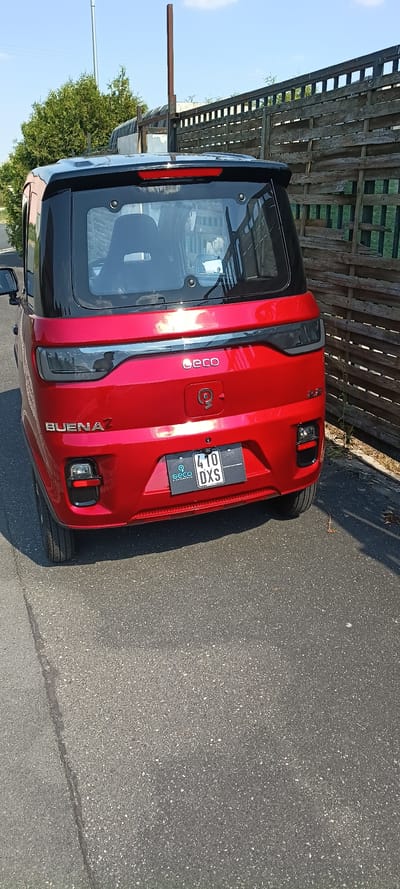 The image shows the rear of a red Geco electric vehicle parked on an asphalt surface. The vehicle has a black roof and black trim around the rear window and taillights. A license plate reading "410 DXS" is visible below the Geco logo. To the right of the vehicle is a weathered wooden fence. In the background, trees and a utility pole are visible under a clear blue sky. The scene appears to be an outdoor area, possibly a residential or commercial property, in Mühlhausen, Germany. There are no people visible in the image.