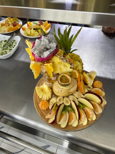 The scene is located within a food preparation or serving area in Chinabad, Uzbekistan. The foreground is dominated by a meticulously arranged, multi-tiered fruit platter presented on a circular wooden board, resting on a brushed stainless steel countertop.
The central fruit platter features carved and sliced fresh fruits. A prominent halved pink dragon fruit, exposing its white flesh speckled with black seeds, is positioned atop a structure composed of cut pineapple pieces and orange segments. A pineapple crown stands upright to the right of the dragon fruit. Beneath this centerpiece, a clear stemmed glass bowl holds additional orange segments and pineapple pieces. At the center of the wooden board, a halved passion fruit, revealing its yellow-orange pulp and black seeds, rests on a stack of thinly sliced, pale yellow fruit, possibly peeled apples or pears. The perimeter of the wooden board is adorned with concentric arrangements of various sliced fruits, including bright orange tangerine segments, pale yellow banana slices, red-skinned apple wedges, and green kiwi slices, often arranged in fan patterns or rows.
To the left and rear of the main platter, on the same stainless steel counter, are three smaller white ceramic plates, each containing an assortment of sliced fruits such as oranges and grapes. Further left, a white oval bowl holds a green salad, possibly an onion and herb mixture, with a metal serving utensil partially submerged.
The environment consists of a reflective stainless steel work surface under bright, uniform artificial lighting. The background features an indistinct, blurred upper wall or overhead fixture and a faint, blurred dark shape on the far right, possibly a person, but without discernible details or actions. No direct human interaction with the fruit is clearly visible in the immediate foreground.