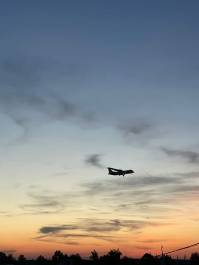 The image displays a twilight sky, likely at sunset, with a prominent dark silhouette of a flying object. The sky exhibits a clear gradient, transitioning from a deep blue-gray on the left to lighter grays and then to warm orange and yellow tones towards the right. Scattered clouds are present, some appearing dark against the lighter sky, others catching the golden light.

The main subject is an angular, dark object, possibly a large kite or a specialized drone, flying in the upper mid-right section of the frame. A thin line, consistent with a kite string or tether, extends upwards from it. Along the far right edge of the image, a dark, silhouetted horizon line reveals the tops of trees and several distant structures, suggesting a town or residential area. No people are visible, and there is no discernible text within the image. The scene conveys a calm and atmospheric moment during dusk or dawn.