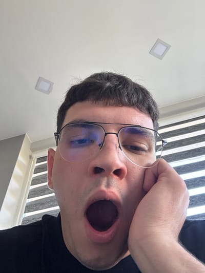 The image is a close-up, eye-level, selfie-style shot of a person's face from the forehead down to their open mouth. The person is wearing aviator-style glasses with thin black frames and clear lenses that reflect a blueish light. Their hair is dark and short. The person's left hand is resting on their right cheek, with fingers splayed. Their mouth is wide open, suggesting they are yawning or speaking. The background consists of a white ceiling with two square, recessed LED lights visible, and a wall with horizontally slatted blinds. The lighting appears to be natural, coming from above and possibly from the right, as indicated by the light reflecting in the glasses and illuminating the right side of the face more strongly. There is no visible indication of the city of Dibich, Bulgaria, in this image.