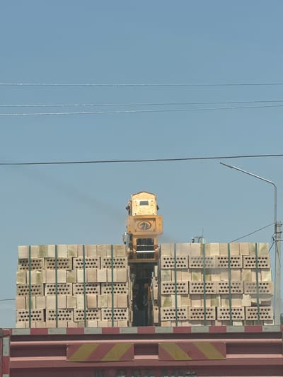 This media file shows a forklift operating near a stack of bricks. The forklift, which is yellow, is positioned in the center of the frame. A large stack of light-colored bricks, bound with green strapping, is to the right of the forklift. The bricks are neatly arranged, with each brick exhibiting multiple circular holes. To the far right of the image is the side of a large, reddish-brown container. There are two hazard stickers on the container: one with diagonal red and white stripes, and another with diagonal yellow and red stripes. The background is a clear, pale blue sky. Three thin, dark cables run vertically from the top to the bottom of the left side of the frame, suggesting the presence of power lines or similar infrastructure. The lighting suggests it is daytime. The scene appears to be an outdoor industrial or logistics area, possibly a port or a construction site. There is no visible text in the image. The orientation of the image is landscape.