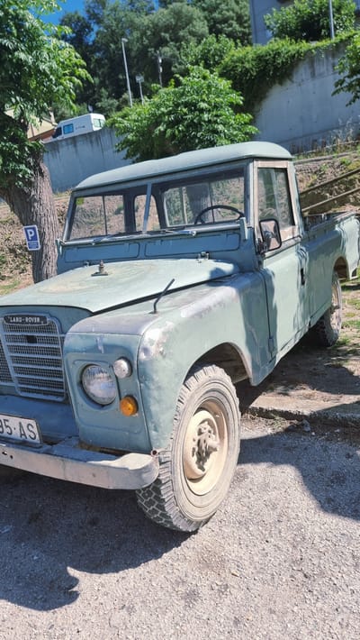 The image shows a light green, vintage Land Rover pickup truck parked on a gravel surface, likely in the city of Castellfollit de la Roca, Spain, given the context. The truck is the primary subject and is viewed from a three-quarter front angle. Its paint is weathered and shows signs of rust and wear, particularly on the front fender and hood.

The truck has a classic design with a prominent grille bearing the "LAND-ROVER" insignia. It features large, round headlights, an amber turn signal, and a chrome bumper. The windshield is split into two sections. A parking sign with a blue "P" is visible on a post to the left of the truck.

The environment appears to be outdoors, with lush green trees and bushes in the background. A concrete wall is visible behind the vegetation. Sunlight casts strong shadows on the ground, indicating a bright day. The overall scene suggests a rural or semi-rural setting, possibly a roadside or parking area. There are no people visible in the image. The image is taken at eye-level with the truck.