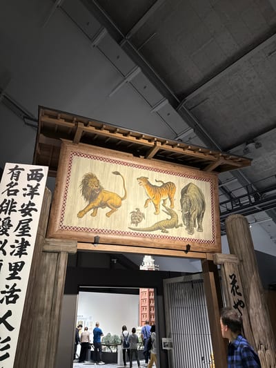 The image shows a museum display in Sumida, Japan. The display features a vertical wooden frame holding a painting depicting an elephant, tiger, crocodile, turtle, and lion, ordered from top to bottom. The painting is surrounded by a brown and white checkered border. Below the painting is a white sign with Japanese characters. To the right of the painting, a man with short grey hair and a blue plaid shirt leans over a wooden beam with black Japanese characters.
In the background, a room displays a white screen with the silhouette of a miniature town. People are walking and standing around the screen.