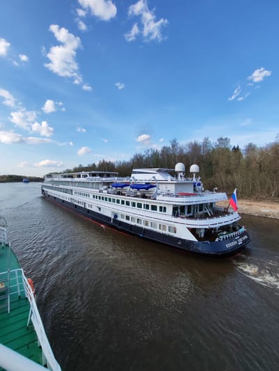 The image shows a river cruise ship, identified as "KNЯЖNА VIKTORIЯ" (Princess Victoria), traveling on a river. The ship is white with a dark hull, multiple decks, and satellite equipment on the upper deck. A Russian flag flies at the stern. The river is dark with visible wake from the ship's movement. The riverbank is visible on the right, with trees lining the shore. The sky is blue with scattered clouds. Part of another ship, green and white, is visible on the left. The image may be taken near Varvarino, Russia, given the style of the ship and landscape, but there is nothing in the image to confirm this city.