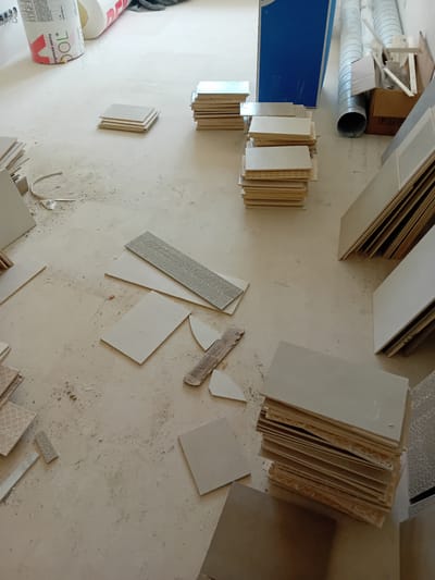 The image depicts a construction site, likely a room interior, with stacks of pale, rectangular tiles laid out on a light-colored, dusty floor. The tiles vary in size and some are stacked neatly, while others are scattered. Some tiles have a textured surface with a raised, repeating pattern, resembling a mosaic or decorative design. Several loose tile pieces and debris are visible on the floor. In the background, a roll of white insulation material with red lettering is partially visible, along with cylindrical tubes, a cardboard box containing construction materials, and a tall blue object. The lighting suggests an indoor environment, possibly under construction or renovation. The image does not contain any people or visible signs that would indicate its specific location within the city of Sloup, Czechia.