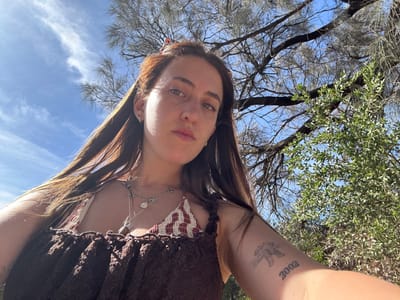 A young woman with long brown hair poses for a selfie under a partly cloudy blue sky. She wears a brown top with a patterned neckline and a silver necklace. Trees with green leaves and bare branches surround her. A tattoo with the year "2002" is visible on her upper arm. The photo is taken from a low angle, looking up at her face.