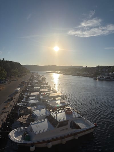 The image depicts a waterfront scene at sunset in the Çanakkale Merkez district of Turkey. A row of small white fishing boats with wooden trim is moored along a concrete pier. The boats are docked side-by-side, with their bows facing towards the left of the frame. One boat in the foreground has the registration "17 D 2094" visible on its hull, along with the word "TURKANA". Another boat further back has "110755" visible. The sun is low on the horizon, casting a warm golden light across the water and the sky, creating reflections on the rippling surface of the strait. Trees and hills line the opposite bank of the water. A few indistinct figures are visible on the walkway beside the moored boats. The overall impression is one of a peaceful evening at a coastal town.