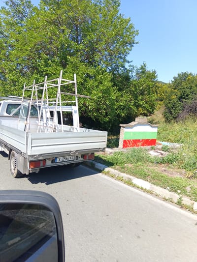 In Golyam Manastir, Bulgaria, a white pickup truck is parked on the side of a road, its bed loaded with window frames and glass. A metal rack is mounted on top of the truck bed, also carrying window frames. In the background, a small structure is painted with the Bulgarian national flag: white on top, green in the middle, and red on the bottom. Lush green trees and vegetation surround the scene under a clear blue sky. A person is visible through the truck's rear window. The road has a concrete curb and the truck's license plate is visible.
