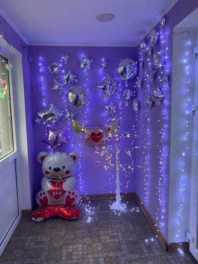 The image shows an indoor space decorated with balloons and fairy lights, possibly in Usolye-Sibirskoye, Russia. Several silver star and round balloons are attached to a purple wall and ceiling. A teddy bear-shaped balloon with the inscription "love you" and a red balloon in the shape of a heart. A decorative white branch with small lights is also present. Fairy lights are strung across the ceiling. The right side of the image shows a tiled wall and flooring, suggesting this area might be a hallway or a transitional space. A window is visible in the lower part of the image.