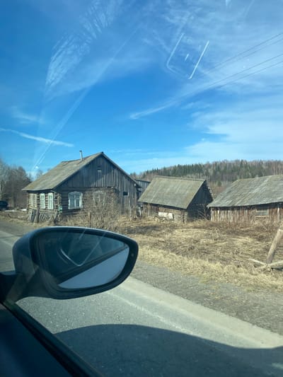 The image shows a view from inside a car, likely driving on a road. The side mirror is visible in the foreground. Outside the car, there are several old, weathered wooden buildings with steeply pitched roofs. The buildings appear to be in a rural or semi-rural setting with dry vegetation in the foreground. In the background, there is a forest and a blue sky with wispy clouds. Overhead power lines are also visible. The location is identified as Okulov, Russia.