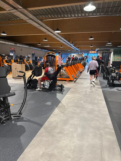 The image shows a gym interior with rows of orange and black Matrix weight machines. People are using the equipment, including one person in red shorts on a leg press. A person in a pink shirt is using a phone. Another individual in a white shirt and black shorts walks by. The gym has a high ceiling with exposed wooden beams, track lighting, and a gray floor with designated areas for machines.