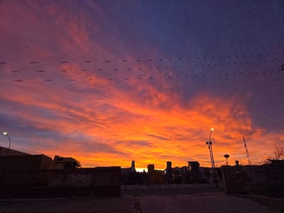 The image captures an urban-industrial scene at sunrise or sunset in Valverde del Majano, Spain. The sky dominates the frame, exhibiting a dramatic gradient from deep violet in the upper third to vibrant orange, red, and yellow hues along the horizon, punctuated by streaky cloud formations. Three dark, parallel horizontal lines, indicative of overhead power lines, traverse the upper portion of the sky. The lower third of the image is characterized by a dark, silhouetted landscape. In the mid-ground, on the left, are several dark, rectangular structures resembling industrial containers or buildings. Partially visible behind these is the dark silhouette of a truck's cab, identifiable by its shape and what appears to be a yellow body. A street lamp on a thin pole, emitting a visible light source, is positioned on the far left. Towards the right, a collection of dark, indistinct shapes suggests additional industrial structures or machinery. Another street lamp, similar in design and illuminated, is situated towards the right-center, mounted on a pole that also features multiple horizontal attachments. Bare tree branches are visible on the far right edge of the frame. The foreground consists of a dark, paved surface. No individuals or discernible activities are present.