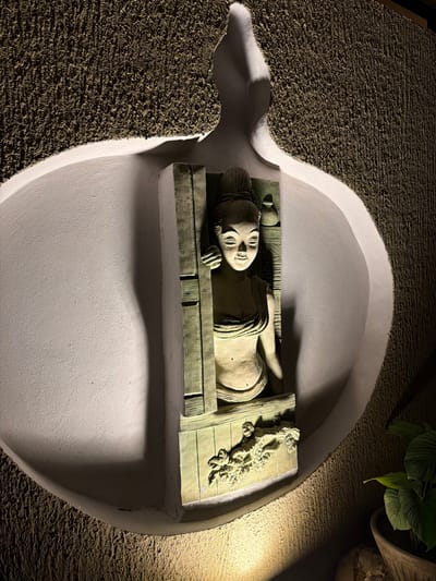 An illuminated sculpture depicting a woman in traditional Thai or Southeast Asian style is prominently displayed within a large, organic-shaped, smooth white niche on a dark, rough-textured wall in Ao Phai, Thailand. The sculpture, primarily in earthy tones with greenish patination, shows a woman with closed eyes and a slight smile, seemingly emerging from a carved frame. Her visible left hand rests on the frame, which features additional decorative carvings of foliage at its base. A small, decorative vessel is positioned on a ledge behind her head to the upper right. The scene is dramatically lit by spotlights from above and below, casting strong shadows within the niche and emphasizing the contrasting textures of the smooth inner surface and the granular outer wall. A potted plant with broad green leaves is partially visible in the lower right foreground.