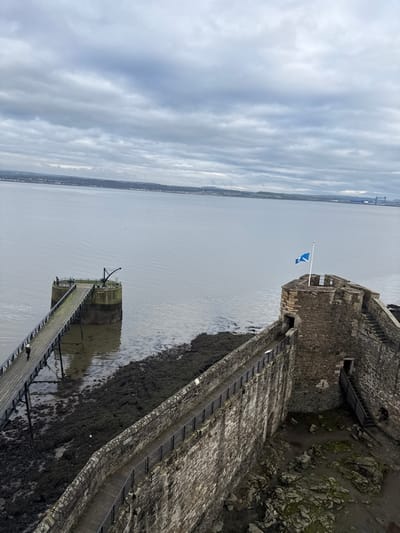 The image depicts an elevated view of Blackness Castle in Blackness, United Kingdom, under a uniformly overcast, gray sky. The foreground features the rugged stone battlements and defensive walls of the castle. A blue Scottish flag, adorned with a white saltire, is flying from a flagpole positioned on the highest point of the castle structure, visible in the upper-right quadrant. A narrow stone walkway, equipped with black metal railings, extends along the top of the castle wall.

To the left of the castle wall, a wooden pier extends into the calm, gray water, connecting to a circular stone platform. A single individual, dressed in light-colored clothing, is observed walking away from this platform along the pier. Two additional, indistinct figures are discernible on the circular platform, near a dark metal crane-like structure. The dark, rocky, and moss-covered shoreline is exposed beneath the pier and along the base of the castle, indicating a low tide.

The midground is dominated by a wide expanse of calm, gray water. In the distant background, a landmass with faint architectural features, including a large bridge structure on the far right horizon, is visible beneath the heavy cloud cover.