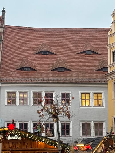 This image captures a multi-story building located in Meissen, Germany, during what appears to be a festive market season. The central structure features a large, red-tiled hip roof with six prominent "eyelid" dormer windows, arranged with one at the top, two below it, and three at the bottom row. Below the roofline, the building's facade is painted a light gray, displaying two visible rows of rectangular windows with reddish-brown frames.
On the upper floor, five windows are visible. The first, third, and fifth windows from the left each contain a white snowflake decoration. The fourth and fifth windows from the left emit a warm yellow interior light. On the partially visible lower floor, four windows display similar frames. The two central lower windows showcase wooden Christmas figurines, including an angel, a Christmas tree, and a Santa Claus figure.
In the foreground, a slender, bare deciduous tree with sparse, reddish-brown leaves stands centrally, partially obscuring the lower facade. Below the tree, the tops of several wooden market stalls are visible. The leftmost stall features a "Handbrot" sign, a large red star, and is adorned with string lights. Another stall to the right also displays string lights and festive decorations. A portion of a yellow building with an ornate baroque-style gable is visible on the far right, adjacent to the central gray building. The sky is overcast and light gray. No individuals are clearly discernible.