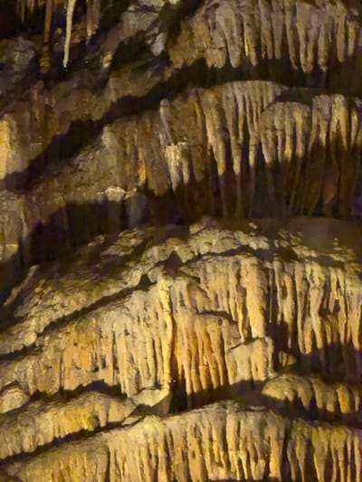 The image depicts the geological formations found inside the Caves of Drach in Manacor, Spain. It features layered rock structures, primarily stalactites and stalagmites, exhibiting a brown and tan color palette. The formations are heavily textured with visible ridges and crevices, illuminated by artificial light, casting shadows.