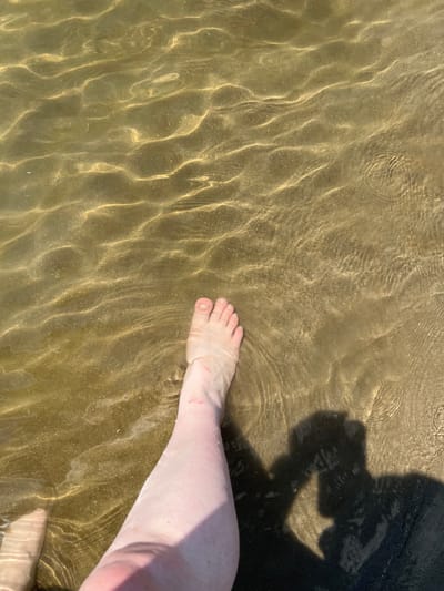 This media file captures a close-up, high-angle shot of a person's leg and foot partially submerged in shallow, clear water. The water creates rippling patterns of light and shadow on the sandy bottom, suggesting a sunny day. The leg, pale in tone, shows some minor abrasions. The foot is bare, with toenails visible.

To the right of the leg, a dark shadow is cast, appearing to be from the person taking the photo or an object they are holding, possibly a camera. The lighting and the shadow suggest this scene is taking place outdoors, likely during daylight hours. The texture of the sand and the clarity of the water are prominent details.

The setting appears to be a natural body of water, such as a beach or a shallow riverbed. Given the location context of Indiya, Moldova, this could be a local river, lake, or a part of the Black Sea coast. No text is visible in the image. The overall impression is one of a relaxed, leisurely moment spent in or near water.