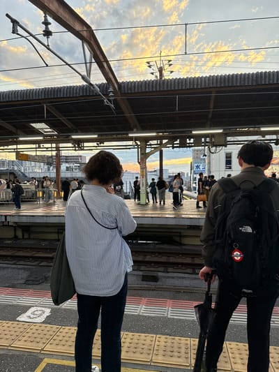 A rear view shows a woman standing on a train platform in Funabashi, Japan. She has dark, curly hair and is wearing a blue and white striped button-down shirt and dark plaid pants. A dark green bag is slung over her left shoulder. Beside her and slightly in front, a person wearing a dark jacket and a black backpack with "The North Face" logo is holding a closed black umbrella. Both individuals appear to be waiting for a train, as they are positioned on the platform overlooking the tracks. In the background, other passengers are visible on the platform, and the sky shows a dramatic sunset with orange and yellow hues. The platform has yellow tactile paving and red and white striped safety lines. Overhead, electrical wires and the corrugated metal roof of the station are visible.