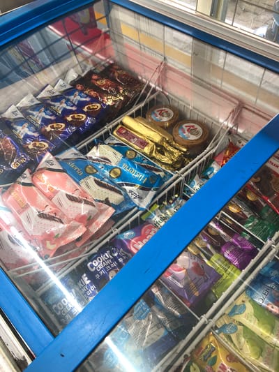 The image shows an open freezer chest filled with various ice cream products, seen from a high-angle perspective. The freezer has a blue trim around the lid and a white metal grid that separates the ice cream. Several rows of ice cream bars are visible, some in wrappers featuring cartoon characters, and others with more generic branding. Some of the ice cream wrappers are translucent, allowing a glimpse of the product inside. Two small round containers with a "K100" logo are also visible. The ice cream is tightly packed, indicating a retail or convenience store setting. The lighting suggests an indoor environment. The context of Putat, Indonesia, implies this is likely a shop or minimarket in that location.