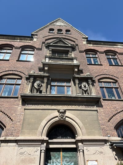 The image shows the facade of a brick building in Lund, Sweden. The building features arched windows and decorative lion sculptures on the upper levels. Below these, the words "AKTIEBOLAGET SYD SVENSKA BANKEN" are inscribed. The main entrance below is an arched doorway with ornate metalwork. The sky is clear and blue.