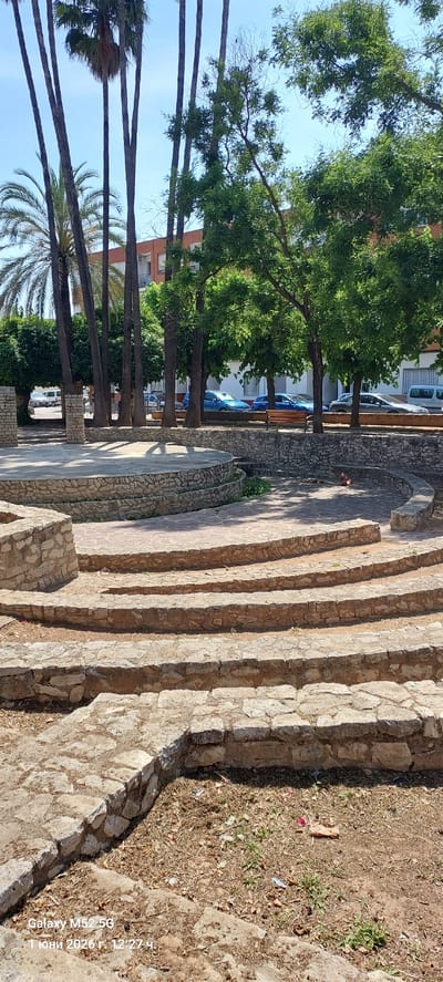 The image shows stone steps leading down to a park area, seemingly in Canals, Spain, as indicated by the date and time stamp "1 июня 2026 г. 12:27 ч."  Palm trees and other trees are visible on the left, and a building with parked cars can be seen in the background. The steps and surrounding stone structures appear aged, and the area adjacent to the steps is covered with dry vegetation.  A person is visible on the stairs.