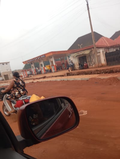 The image depicts a street scene in Imilike, Nigeria, with a focus on a woman riding a motorbike in the foreground. The woman is wearing a floral-patterned dress and appears to be carrying yellow containers on the back of her motorbike. The motorbike is white and red. In the background, a gas station is visible with orange and white pumps and an awning. Several people are present at the gas station, and a truck is parked nearby. The environment appears to be dusty and unpaved, with reddish-brown earth covering the ground. A utility pole with wires is also present. The scene is viewed from the perspective of someone in a vehicle, as a car's side mirror is visible in the lower right corner of the image, reflecting a person wearing a black shirt with white lettering. The sky is overcast and appears bright.