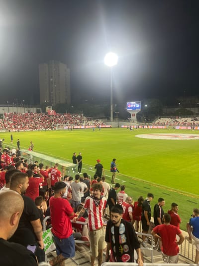 The image depicts a night-time soccer match or event in a stadium located in Nof HaGalil, Israel. The perspective is from the stands, looking towards the field and the opposite side of the stadium.

The environment is illuminated by two prominent stadium floodlights, one on the left and one on the right, casting a strong glare and creating a bright green field. The sky above is dark, indicating night. In the far background, a multi-story residential building is visible.

The stands on the left and right are densely populated with spectators, predominantly male, many wearing red and white apparel, suggesting support for a team. A large digital scoreboard is visible in the mid-ground, displaying a "0:0" score and "00:00" time, with team logos and the text "NOF HAGALIL" below.

On the illuminated green field, several small figures, presumably players or coaching staff, are dispersed. Some individuals are wearing blue uniforms, others dark attire.

In the immediate foreground, a group of male spectators stands on concrete stadium steps. Many are wearing red shirts. Specific individuals include: a man with a shaved head and a black t-shirt in the lower left; a man in a red t-shirt and light blue cut-off jeans with visible tattoos on his lower legs; and a man in a red and white striped shirt with a patterned bandana around his neck, looking towards the camera. Another man in a black t-shirt with a graphic design is positioned in the lower right. Most individuals are observing the field, with some appearing to be in motion or conversing.