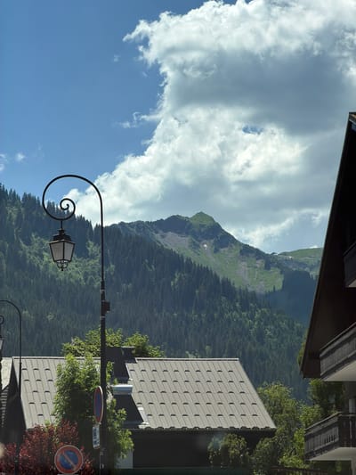 This is a vertical, eye-level medium shot photograph taken in Châtel, France. The scene features a mountain landscape with a heavily treed slope that rises towards a rocky peak. The sky is partly cloudy, with large, fluffy white clouds against a bright blue background. In the foreground, a black wrought-iron lamppost with a scrolled arm and a lantern is visible, silhouetted against the sky and mountains. To the right of the lamppost, a modern building with a gray, tiled facade is prominent. Two circular traffic signs are also visible on posts near the building. The lighting suggests it is daytime, possibly late morning or early afternoon, with a bright sun illuminating the scene. The overall mood is serene and picturesque, showcasing the natural beauty of the mountains juxtaposed with modern architecture. No people are visible in the image. The traffic signs are small and difficult to read clearly, but one appears to indicate a parking restriction.