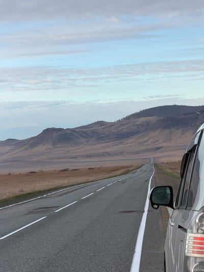 The image shows a gray car parked on the shoulder of a two-lane asphalt road. A white line marks the road's edge. The landscape features a large, sloping hill or mountain covered in dry, brown vegetation. The sky is overcast with light blue and gray clouds. The location is rural, consistent with an area outside a city, and unlikely to be within Aeshka, Russia. No people are visible.