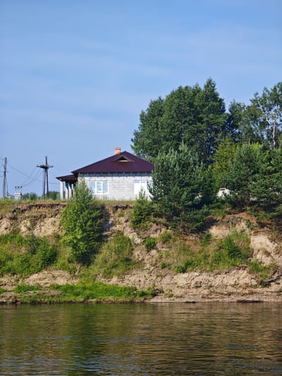 In the city of Galanino, Russia, a construction site is visible on a steep, sandy riverbank. A partially built house, constructed of concrete blocks, stands at the top of the bank, featuring a dark brown metal roof and white-framed windows covered with protective film. Overhead power lines and utility poles are present on the left side of the image. Dense green trees, including pines and deciduous varieties, occupy the upper portion of the riverbank, partially obscuring the house and extending towards the right edge of the frame. The river in the foreground reflects the green foliage and the blue sky, with ripples on its surface. The overall scene depicts a rural or semi-rural setting with ongoing construction activity.