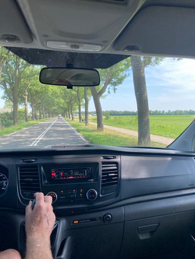 The image shows the interior of a vehicle, likely a van or truck, as seen from the driver's perspective. The focus is on the dashboard and the view through the windshield. The dashboard is made of dark plastic and features a car stereo, air vents, and a hand reaching towards what appears to be a gear shift. The stereo displays "JVC 32 STEP" in red lettering. To the right of the stereo, an orange warning light is visible, along with text reading "PASSENGER AIRBAG." Below that, a circular 12V power outlet is present.

Through the windshield, a road lined with trees stretches into the distance, suggesting a rural or suburban setting. The trees are tall and mature, with green foliage, and the road has white lines marking the lanes. The sky is blue with some clouds, indicating daytime with fair weather. The scene is captured during daylight hours, likely mid-morning or afternoon, given the brightness of the natural light. The overall impression is that of a journey in progress through a scenic, tree-lined avenue. Visible text includes "12V", "AIRBAG", "PASSENGER AIRBAG", "DEAR", "JVC", "32 STEP", and numbers on a dial that seem to indicate speed, such as "120", "140", and "200". The image is oriented vertically, suggesting it was taken with a phone held in portrait mode, or has been rotated.

---
**Note:** The provided OCR results contain some elements that might not be perfectly aligned with the visual content. For example, "DEAR" and "Q" don't appear as distinct words or symbols in the visible parts of the dashboard in the image. The numbers "120", "140", and "200" likely refer to a speedometer dial, which is partially visible at the bottom of the image. "0" and "0-" are also visible on this dial. The "12V" is clearly a power outlet. The "AIRBAG" and "PASSENGER AIRBAG" text is also correctly identified.