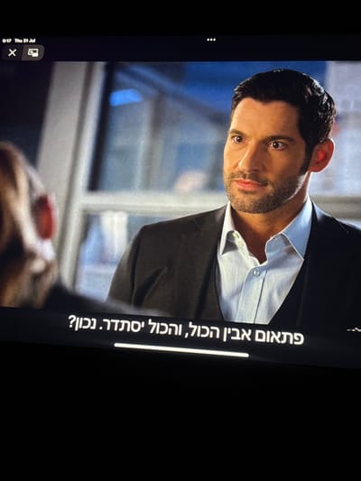 The image displays a digital screen captured at an angle, showing a video scene from the television series "Lucifer." The central subject is a male individual, identified as Tom Ellis portraying Lucifer Morningstar, viewed from the chest up. He has dark hair, a short beard, and is wearing a dark suit jacket, dark vest, and light blue collared shirt. His gaze is directed towards the left foreground, where the blurred back and right shoulder of a second, unidentified individual are partially visible, suggesting an interaction. The background is an out-of-focus indoor setting with indistinct architectural elements and subtle blue lighting. Overlaid on the screen content are user interface elements: "0:17 Thu 31 Jul" in the upper left corner, along with "X" and casting icons. Hebrew subtitles are present at the bottom of the screen, reading "פתאום אביו הכול, והכול יסתדר. נכון?" (Suddenly his father, and everything will work out. Right?). This photograph of the screen was taken in Kafr Bara, Israel.