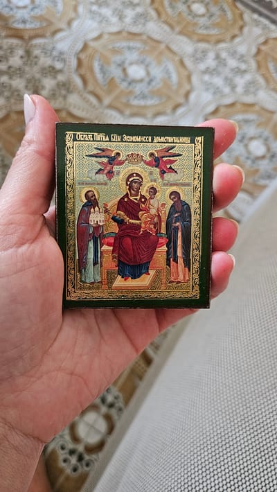 An antique religious icon is held in a person's hand. The icon is square and depicts the Virgin Mary holding the Christ Child, flanked by two saints and two angels. The background of the icon is gold. The icon is framed by a dark green border. The person's hand is holding the icon with their thumb and index finger on the left side, and their middle, ring, and pinky fingers on the right side. The background of the image is a patterned textile. There is no discernible location in the city of Mondolfo, Italy.