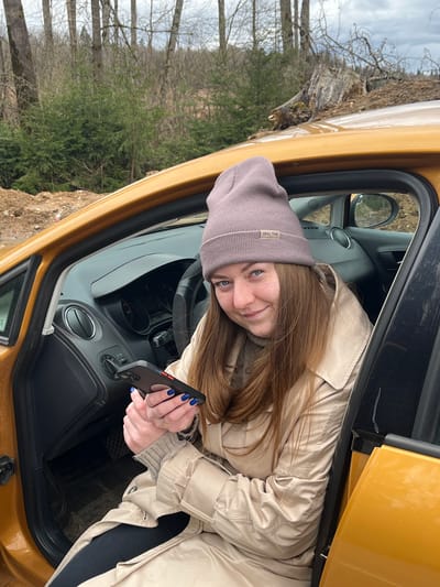 A woman with long brown hair and blue eyes, wearing a gray beanie and beige trench coat, is seated in the driver's seat of a gold-colored car. She is holding a black cell phone in her hands, her fingernails painted blue. The car door is open, and the exterior reveals a wooded area with bare trees and some green foliage. The scene appears to be outdoors, with an overcast sky. The location is stated to be Ivlevo, Russia, though this cannot be confirmed from the image.