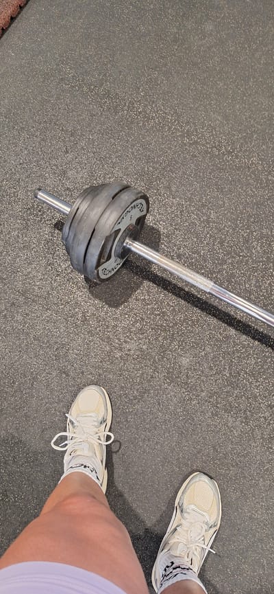 An overhead shot captures a person's lower legs and feet next to a loaded barbell, set against a dark grey, speckled rubber gym floor. The person wears light-colored athletic shoes with white laces and white socks adorned with black patterns. Their bare calves and knees are visible, suggesting they are wearing shorts or athletic attire.
To the left, a shiny silver barbell lies diagonally across the frame. One end of the barbell is fitted with several black weight plates. Clearly visible on the topmost weight plate is the brand name "TECHNOGYM" and the number "5", likely indicating its weight (e.g., 5 kg). The barbell casts a noticeable shadow on the textured floor. The scene implies an indoor setting, most likely a gym or fitness facility in Santa Cruz De Tenerife, Spain, with the person seemingly paused during a workout. No specific time of day or emotions are discernible.