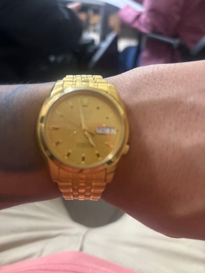 The image displays a close-up, slightly out-of-focus view of a person's left wrist adorned with a gold-toned wristwatch featuring a matching metal link bracelet. The watch face is gold-toned with gold-toned hour markers, three hands, and a day/date complication at the 3 o'clock position, showing "MON 20" or similar text. The skin on the wrist and lower arm appears light-to-medium in complexion, with some visible hair and a faded tattoo on the left side of the arm. The person's lower torso is partially visible, showing light-colored pants and a pink garment underneath. The background is heavily blurred, indicating an indoor environment, with indistinct elements including dark clothing on the left, a light-colored surface or object directly behind the watch, and a maroon/pink garment and dark blue object on the right. There are no visual cues within the image to confirm the specific location within the city of Adrogué, Argentina.